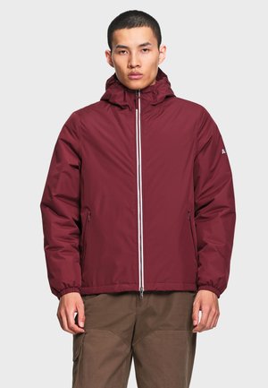 Jeune homme portant une veste à capuche bordeaux avec une fermeture éclair devant et un pantalon marron, debout devant un fond clair uni.