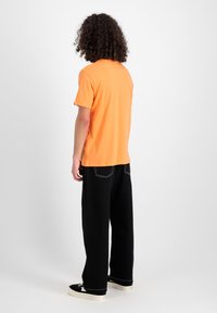 Oranges T-Shirt mit kurzen Ärmeln und geradem Schnitt, kombiniert mit schwarzen weit geschnittenen Hosen mit weißen Stickdetails und schwarzen Sneakers.