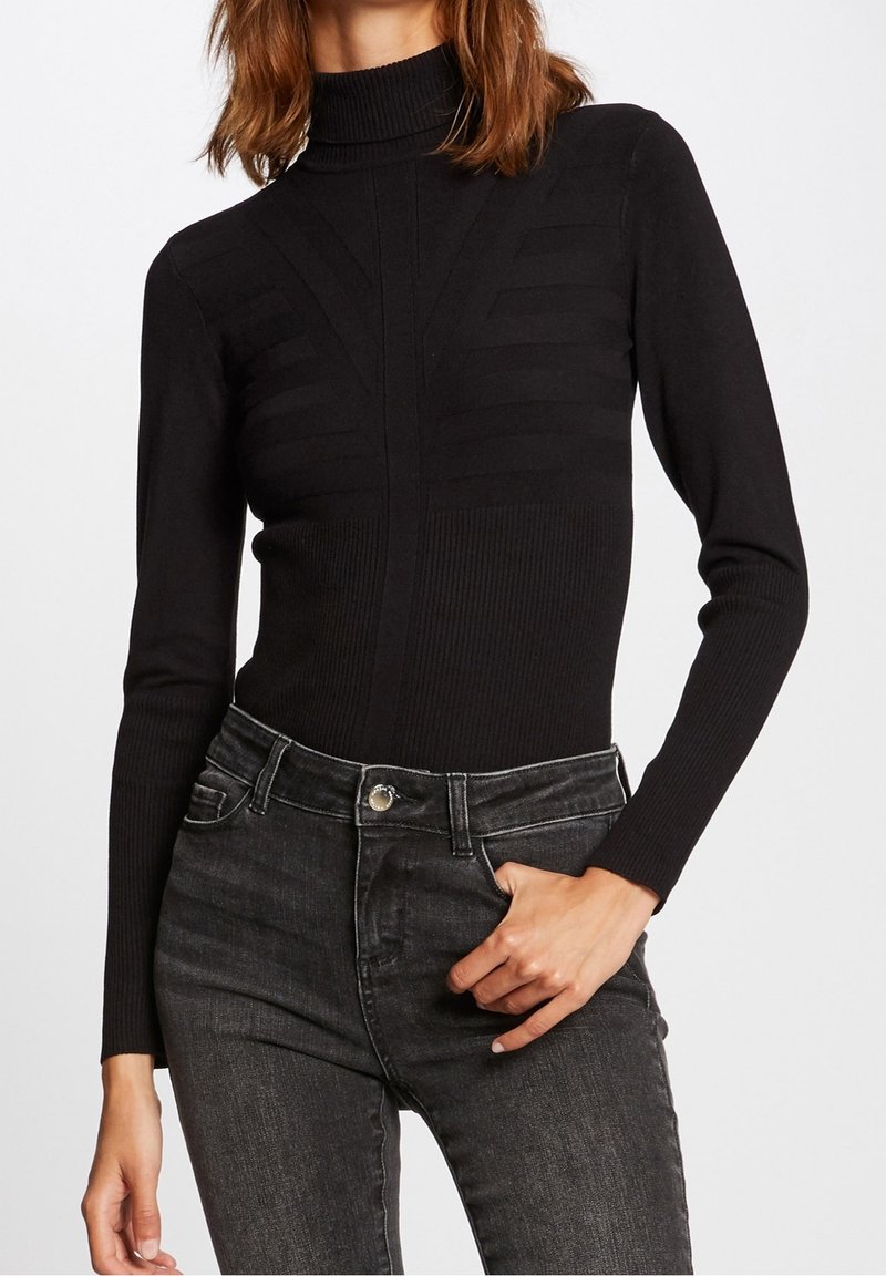 Mujer vestida con un suéter negro de cuello alto ajustado y acanalado con patrones geométricos, combinado con jeans negros desgastados de cintura alta.