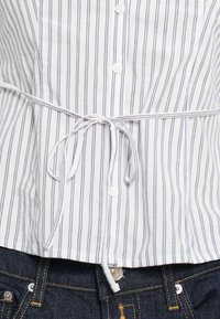 Fehér ing, függőleges kék csíkokkal, vékony övvel a derekán megkötve. Az ing galléros és sima tapintású.