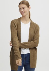 Bruine gebreide cardigan met lange mouwen, open voorkant en textuurstof. Heeft ribbels aan de manchetten en zijkanten zakken. G gedragen over een wit shirt.