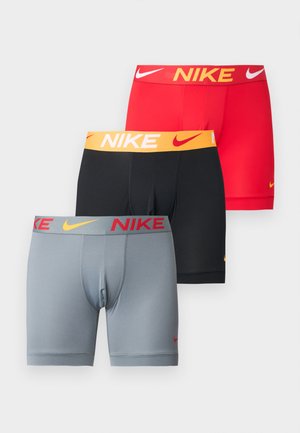 Atletyczny mężczyzna o ciemnej karnacji stojący boso w jasnoszarych bokserkach Nike z pomarańczowymi logo na jednolitym białym tle.