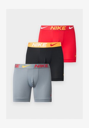 Atletyczny mężczyzna o ciemnej karnacji stojący boso w jasnoszarych bokserkach Nike z pomarańczowymi logo na jednolitym białym tle.