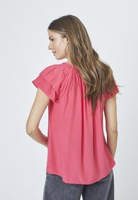 Blusa rosa a maniche corte con scollo elasticizzato, texture liscia e orlo curvo, abbinata a jeans grigi.