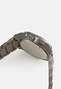 Orologio in metallo scuro con cassa esagonale, anelli del bracciale texture e pulsanti in rilievo. Il retro è in acciaio inossidabile con dettagli incisi.