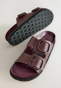 Sandales en synthétique marron avec deux boucles réglables, une semelle en caoutchouc noir texturé et un lit de pied maroon lisse.