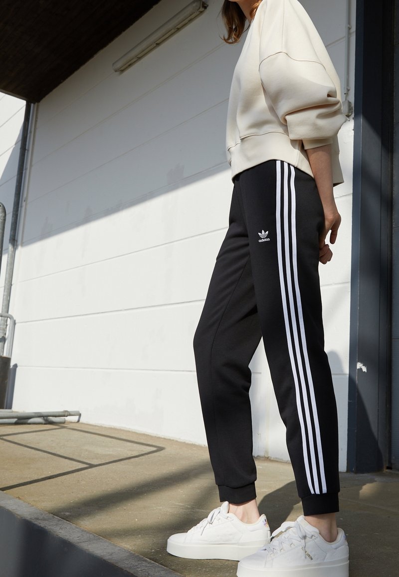 Svarta träningsbyxor med vit tre-stripes-design, parat med en krämfärgad sweatshirt. Modellen har på sig vita sneakers. Avslappnad athleisure-stil.