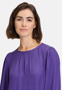 Lilla bluse med lange puffærmer og rund halsudskæring, der har plisserede detaljer foran og et glat, let stof.