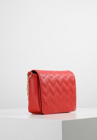 Borsa a tracolla in pelle trapuntata rossa con patta magnetica, tracolla a catena e motivo a zigzag sulla parte anteriore, caratterizzata da una texture liscia.