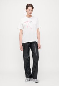 Vit t-shirt med en rosa bandgrafik och text; ihopparad med svarta fuskläderbyxor och gråa träningsskor. Enkel design, avslappnad passform.