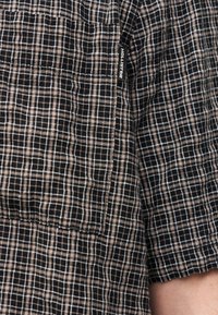 Κοντινό πλάνο ενός μανικιού πουκάμισου Hollister με καρό μοτίβο σε μαύρο και μπεζ χρώμα και με ορατή ετικέτα της μάρκας ραμμένη στη ραφή του πλαισίου.