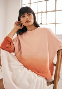 Oversized bluza w pastelowym różu z pomarańczowym gradientem, wykonana z miękkiego materiału. Posiada okrągły dekolt i ściągacze przy mankietach. Biała spódnica dodaje kontrastu.