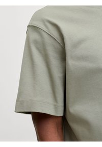 Κοντινό πλάνο ενός ατόμου που φοράει μια κοντομάνικη πουκάμισα σε ανοιχτό χακί πράσινο χρώμα, εμφανίζοντας τις λεπτομέρειες της ραφής στο μπράτσο και τον ώμο.