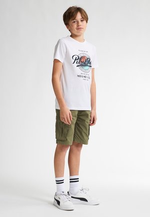T-shirt bianco con stampa grafica, pantaloni cargo verdi, sneakers bianche con dettagli neri e calze bianche con righe nere. Design semplice, vestibilità casual.