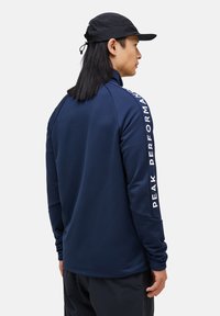 Marineblå sportsbluse med lange ærmer, der har "PEAK PERFORMANCE" trykt i hvidt ned ad højre ærme. Glat stof og moderne snit.
