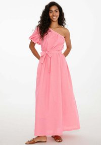 Ružová maxi šaty na jedno rameno s volánovým rukávom, zúženým pásom a splývavou látkou. Nosené s jednoduchými hnedými sandálmi.
