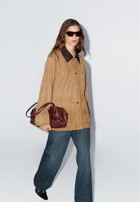 Giacca in suede marrone con colletto nero, grandi bottoni frontali, abbinata a pantaloni a zampa d'elefante in denim scuro e una borsa bordeaux. Design senza cuciture.