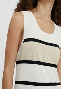 Top in maglia senza maniche con ampie righe orizzontali oro e nere indossato da una donna con capelli ricci, corto sulle spalle e sul collo.