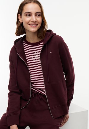 Giovane donna sorridente che indossa una felpa bordeaux con zip, pantaloni abbinati e una maglietta rosa e bordeaux a righe con un logo.