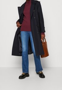 Marinblå trench coat med dubbelknäppt design, vinröd ribbad tröja, blå utsvängda jeans, svarta plattformsskor och brun handväska.