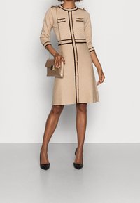 Krämfärgad klänning med strukturerat mönster, svart kant, gulddetaljer och två bröstfickor. Tillsammans med en guldig kuvertväska och svarta klackar.