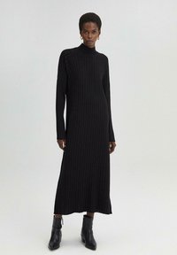 Neulottu musta midi-mekko, jossa on pystysuora ribbineulos, korkea kaulus, pitkät hihat ja sivurei'itykset. Yhdistetty mustiin nilkkabootseihin.