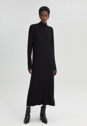 Neulottu musta midi-mekko, jossa on pystysuora ribbineulos, korkea kaulus, pitkät hihat ja sivurei'itykset. Yhdistetty mustiin nilkkabootseihin.