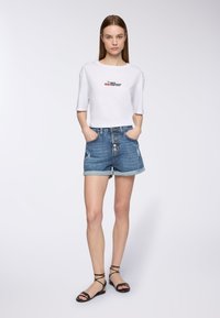 Biała koszula z krótkim rękawem z grafiką czereśniową oraz dżinsowe szorty z podwiniętymi nogawkami i guzikami z przodu. Czarne sandały na paski dopełniają stylizację.