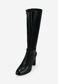 Bota de cuero negro hasta la rodilla con tacón cuadrado y hebilla decorativa alrededor del tobillo, expuesta sobre un fondo blanco.
