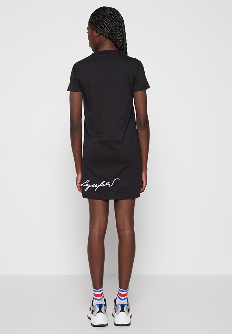 Robe t-shirt noire à manches courtes, ornée de texte en script blanc au niveau de l'ourlet. Stylée avec des chaussettes de sport multicolores et des baskets.