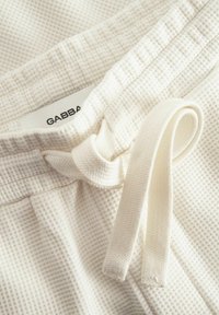 Vitt vävt tyg med en texturerad mönster; har ett dragsnöre och en etikett som säger "GABBA" nära midjan.