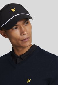 Black cap with white trim and yellow eagle logo, worn with a navy sweater and black collared shirt, featuring a V-neck design.