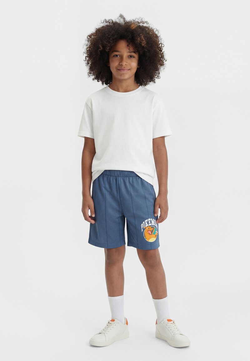 Dziecko z kręconymi włosami nosi białą koszulkę, niebieskie spodenki z motywem Pokémon, białe skarpetki i białe trampki, stojąc na białym tle.