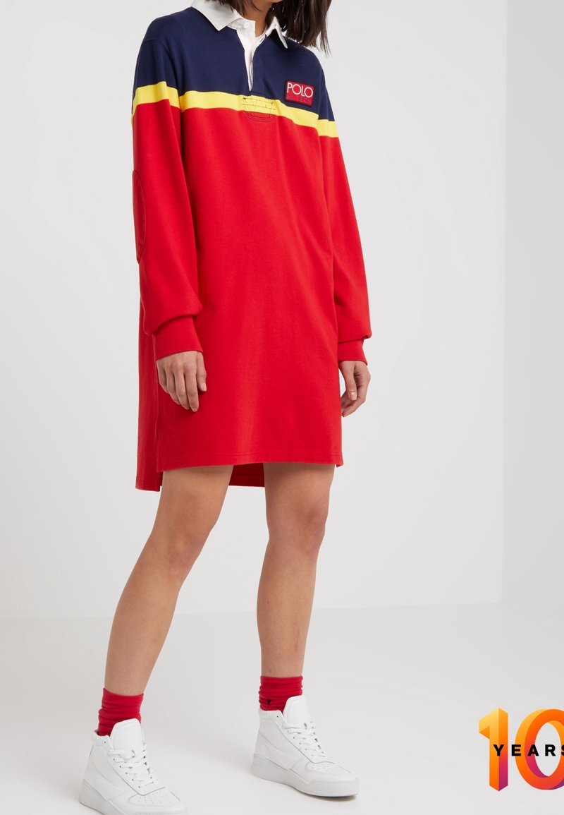 Röd och marinblå rugbyinspirerad klänning med långa ärmar, vit krage och gul rand över bröstet. Har sidosprund och ribbade manschetter.