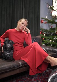 Röd stickad klänning med långa ärmar och utsvängd nederdel, tillsammans med en svart mops som sitter på en brun lädersoffa mot gröna gardiner.