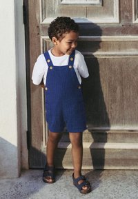 Blue corduroy overalls with wooden buttons, paired with a white t-shirt and dark sandals. Standing against a wooden door.