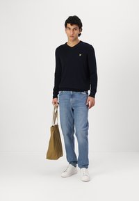 Jersey de pico azul marino, jeans de color azul claro, zapatillas blancas, sosteniendo una bolsa de tote color beige. El jersey presenta un pequeño logo amarillo en el pecho.