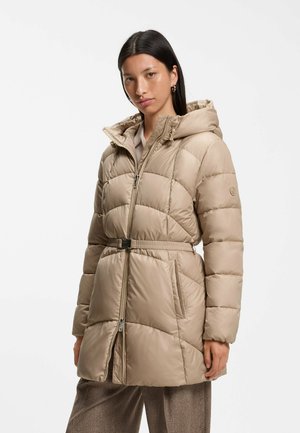 Donna che indossa un cappotto imbottito trapuntato beige con cappuccio e cintura, in piedi su uno sfondo bianco semplice.