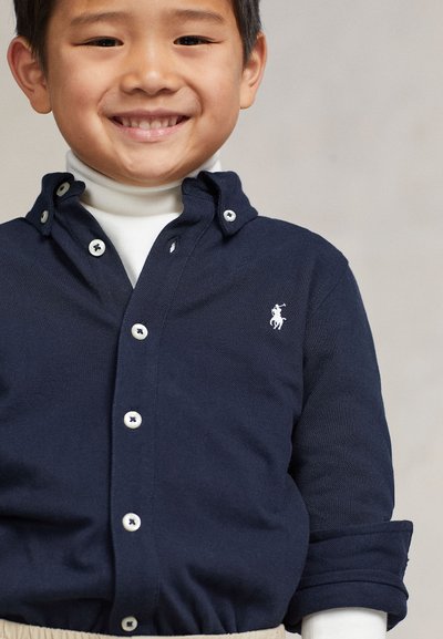 Jeune garçon souriant portant une chemise bleu marine à boutons avec un logo polo blanc sur un col roulé blanc, pantalon beige visible.