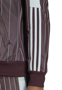 Bordeauxroter Sportjacke mit vertikalen weißen Streifen, gerippten Bündchen und seitlichen Reißverschlusstaschen; mit AEROREADY-Logo auf der unteren linken Seite.