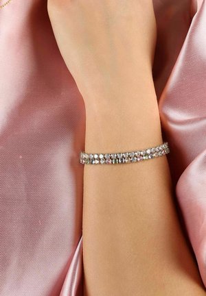 Silberarmband geschmückt mit mehreren funkelnden Steinen in verschiedenen Farben. Am Handgelenk getragen vor einem sanften rosa Satin-Hintergrund.