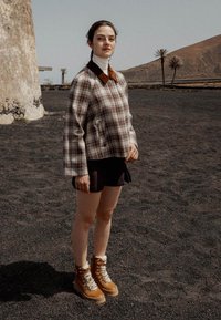 Chaqueta de cuadros en marrón claro, blanco y marrón oscuro, con un cuello marrón; pantalones cortos negros; botas marrones con forro blanco y cordones.