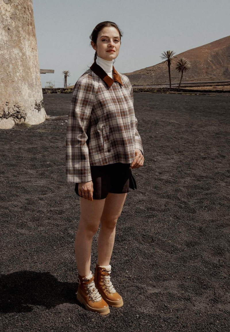 Chaqueta de cuadros en marrón claro, blanco y marrón oscuro, con un cuello marrón; pantalones cortos negros; botas marrones con forro blanco y cordones.