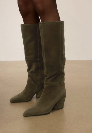 Bottes hautes jusqu'au genou en daim vert olive avec des bouts pointus et des talons épais, portées sur des jambes nues sur un fond uni.
