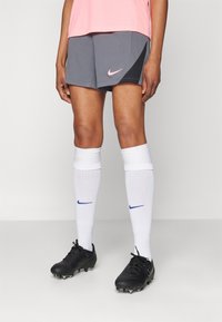 Nike atletske kratke hlače u sivoj boji s ružičastim logotipom, uparene s bijelim čarapama do koljena koje imaju plavi logotip, te crnim kopačkama s vidljivim kopačkama.
