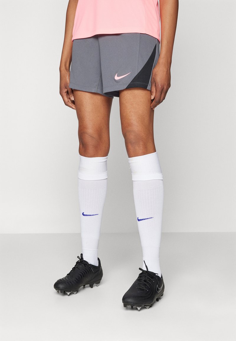 Nike atletske kratke hlače u sivoj boji s ružičastim logotipom, uparene s bijelim čarapama do koljena koje imaju plavi logotip, te crnim kopačkama s vidljivim kopačkama.