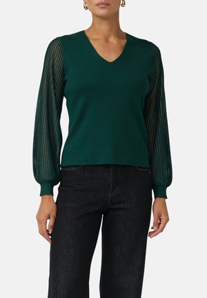 Femme portant un pull en V vert foncé avec des manches longues en voile rayé et un pantalon noir, se tenant devant un fond uni.