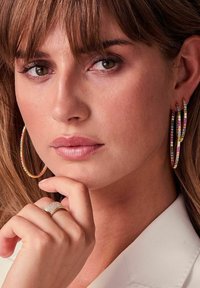 Orecchini a cerchio dorati con pietre multicolori e un anello dorato texturizzato con piccoli diamanti. Primo piano del viso e della mano.