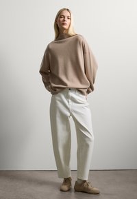 Beige gestrickter Oversize-Pullover mit hohem Kragen, kombiniert mit weißen, locker sitzenden Hosen und hellbraunen Sneakers. Sanfte Texturen, lässiges Design.