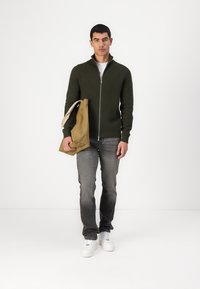 Grønn zip-up genser, grå bukser, hvite sneakers, og en brun skulderveske. Genseren har høy krage og ribbede mansjetter.